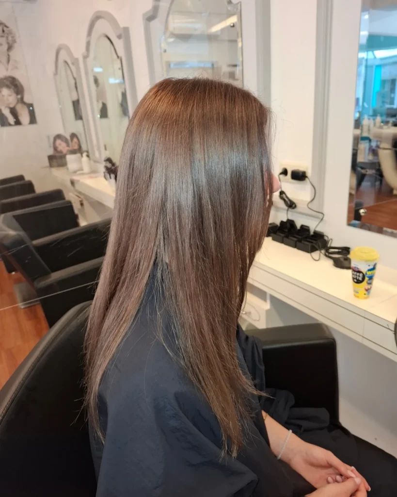 Back view of a person with long straight brown hair seated in a salon chair, with styling tools and products visible on a counter in the background.