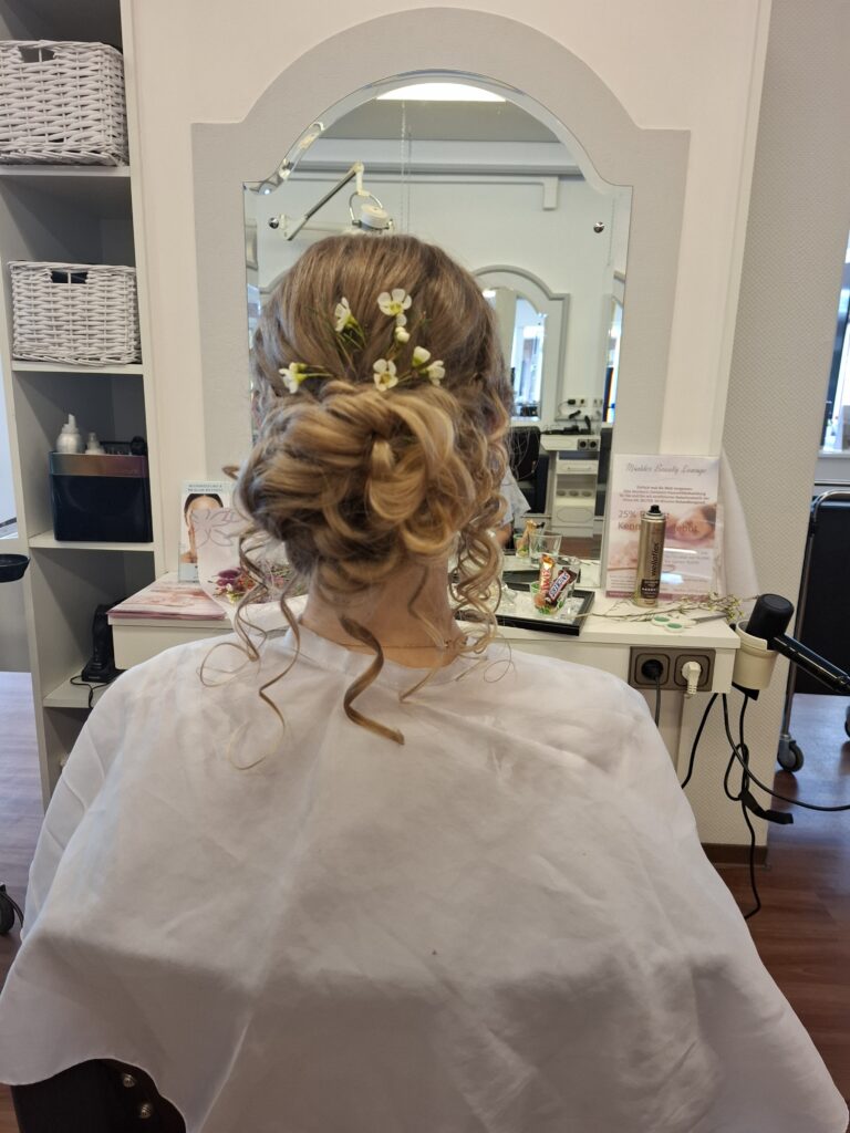 A person seated in a salon with hair styled in an elegant updo adorned with small yellow flowers, wearing a white cape, with salon equipment in the background.