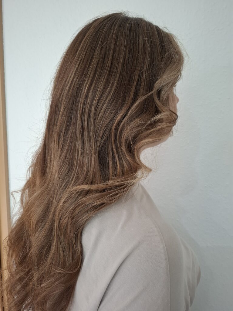 Side view of a person with long, wavy brown hair wearing a white top, looking slightly downward.