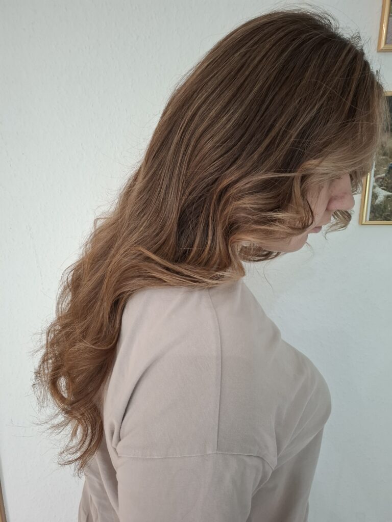 Back view of a person with long, wavy brown hair wearing a light-colored top, against a plain white background.