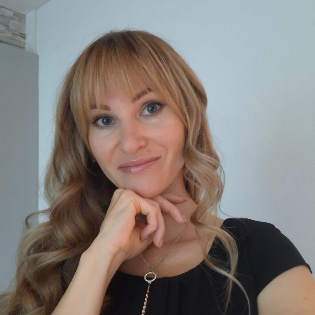 A person with long, wavy blonde hair wearing a black top and a delicate necklace with a circular pendant, resting their chin on their hand, against a plain indoor background.